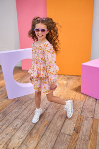 Child in a floral top and skirt wearing sunglasses posing in front of colorful geometric shapes.